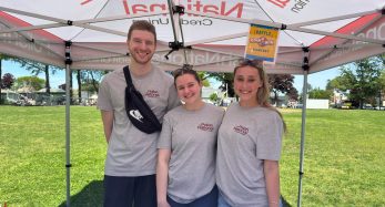 Three smiling volunteers under a promotional tent at a park event, wearing gray shirts with a Polish National Credit Union logo.