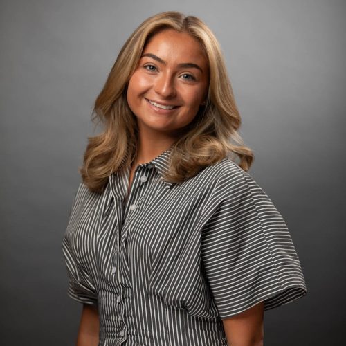 Smiling woman with shoulder-length blonde hair in a striped button-down shirt against a gray studio backdrop.